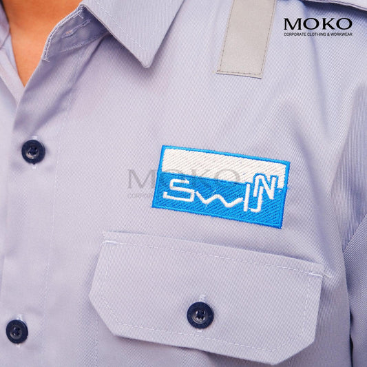 Close-up of a light blue corporate work shirt with a blue embroidered name patch and pocket.